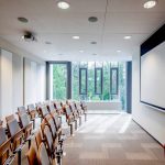 Sala wykładowa ze składanymi krzesełkami naprzeciw tablicy do wyświetlania informacji.